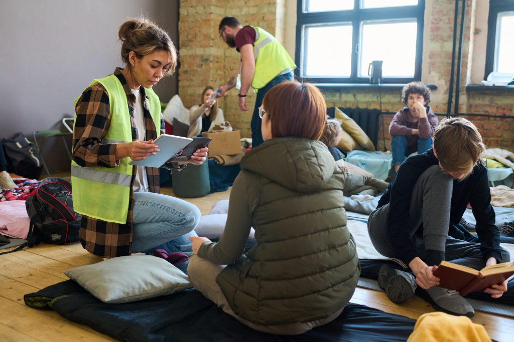 Female volunteer with mobile gadgets sitting on squats in front of refugee sitting on her sleeping place in temporary shelter